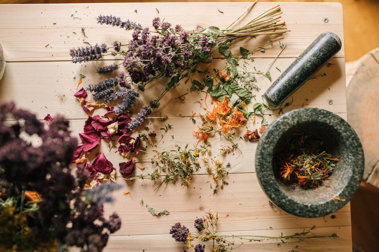 Pourquoi-nous-choisir ? Flat lay of dried herbs and flowers with a stone mortar and pestle on a rustic wooden table.